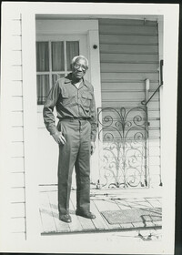 Photograph Philip Simmons standing next to window grate on his porch.